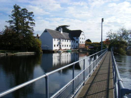 Photo over a river to a large white painted building and a lock. Taken from a footbridge over a weir.