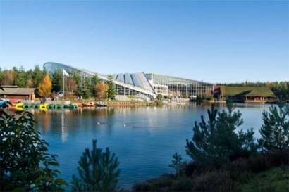 Photo of the main building at CenterParcs Whinfell Forest, a glass building with a sweeping curved roof. Taken from across a blue lake on a day with a clear blue sky.